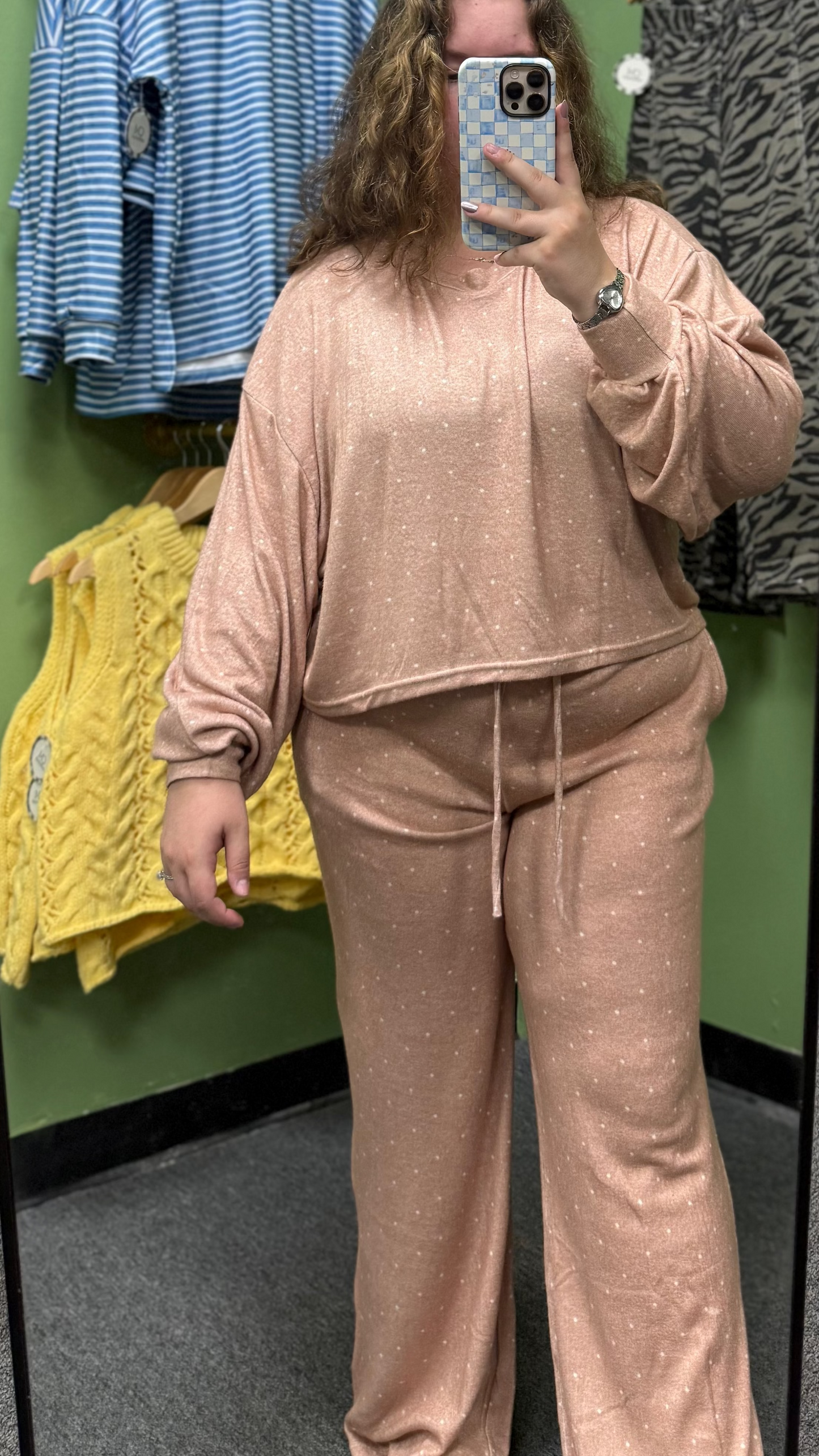 A person taking a mirror selfie in a clothing store, wearing a matching blush pink lounge set consisting of a cropped long-sleeve top and wide-leg pants. The background shows a green arched wall with other clothes hanging on racks.