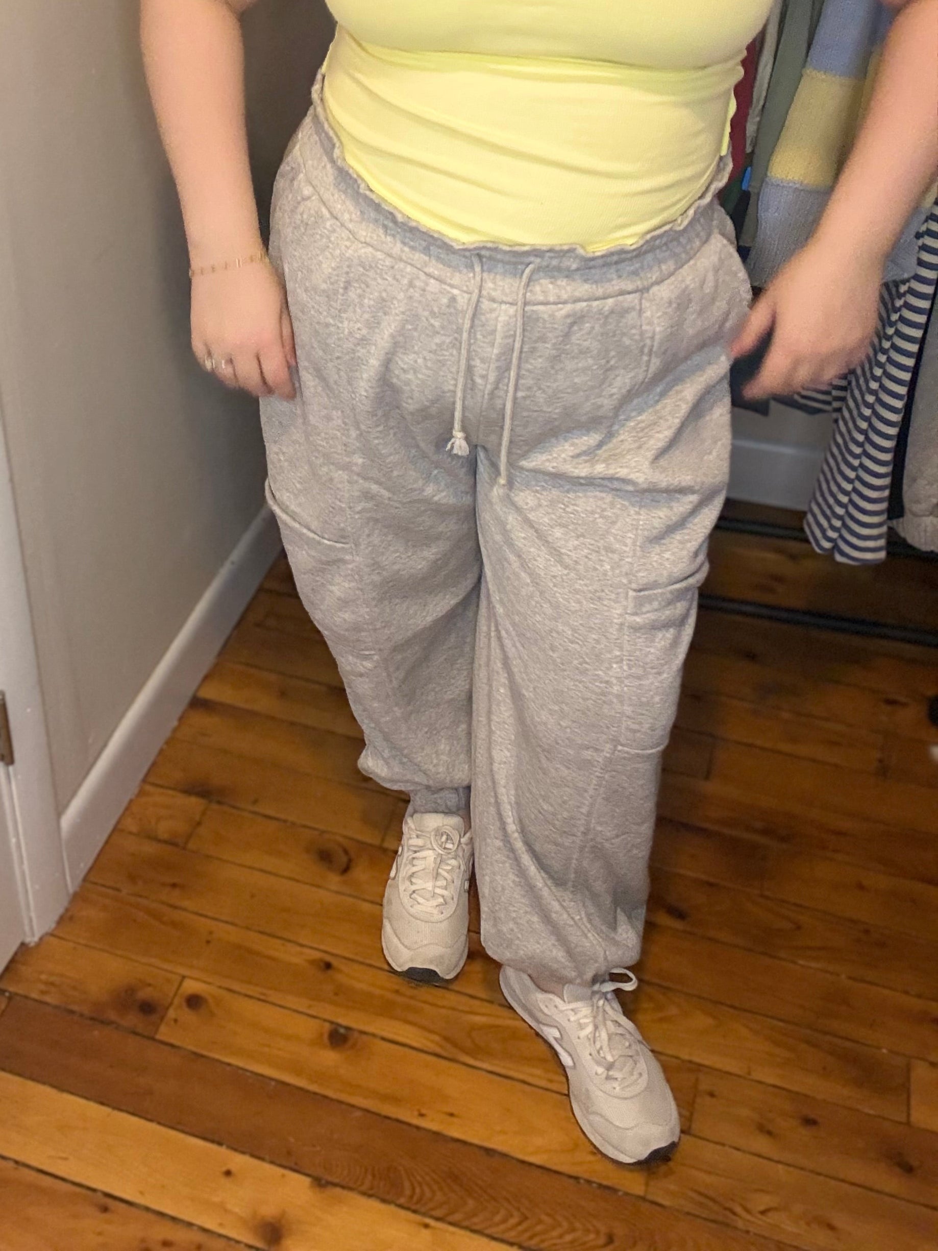 Person wearing a yellow tank top and gray sweatpants standing in a room with wooden flooring.