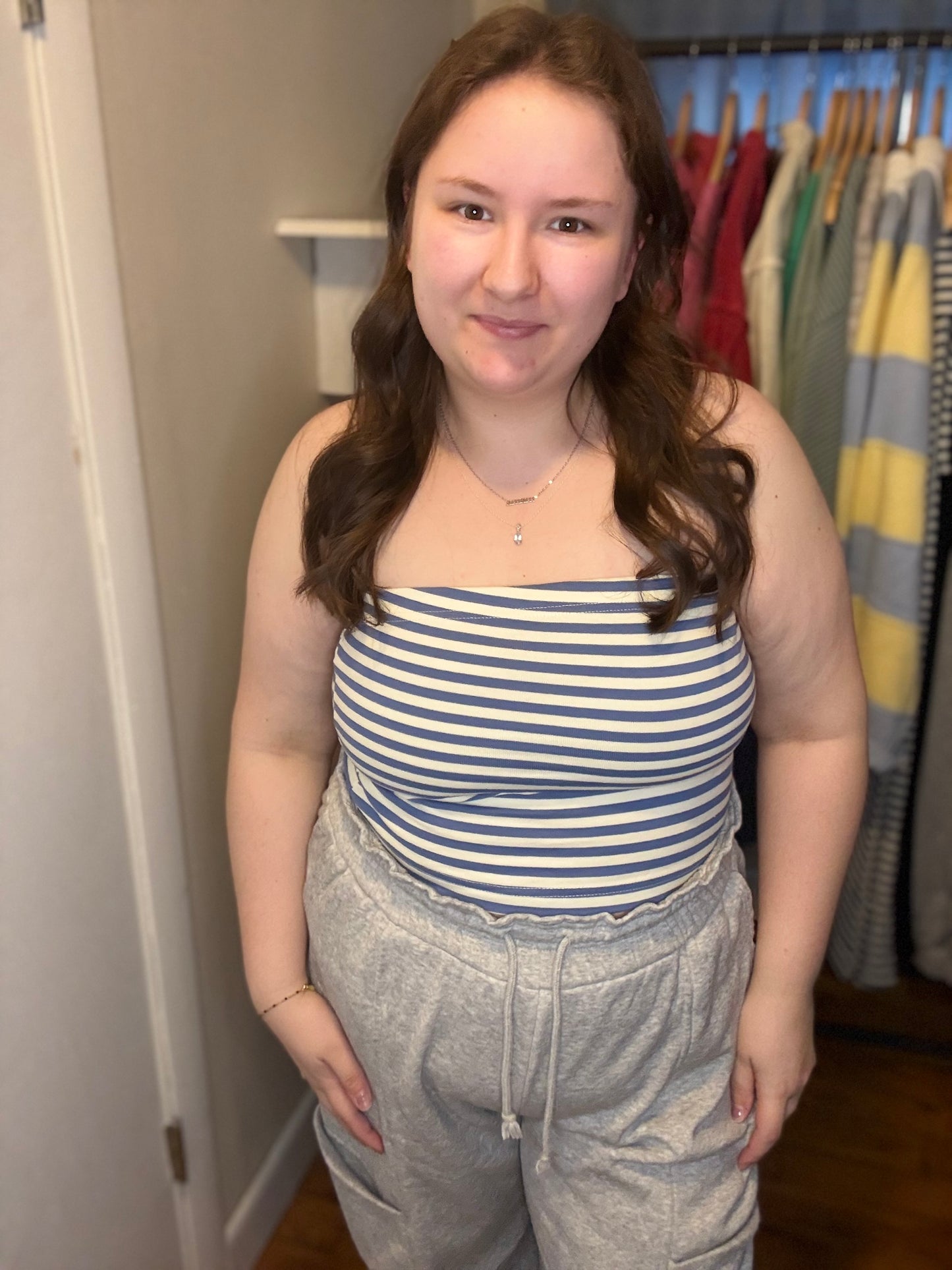 Woman wearing a striped tube top and gray sweatpants in a room with clothes on hangers.