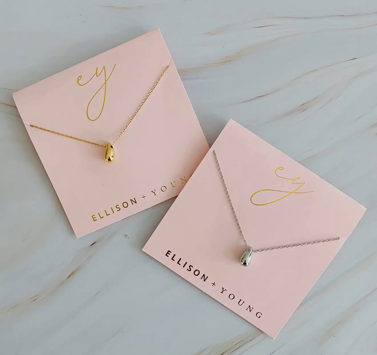 Two necklaces on pink cards with 'ELLISON + YOUNG' branding on a light wooden surface.