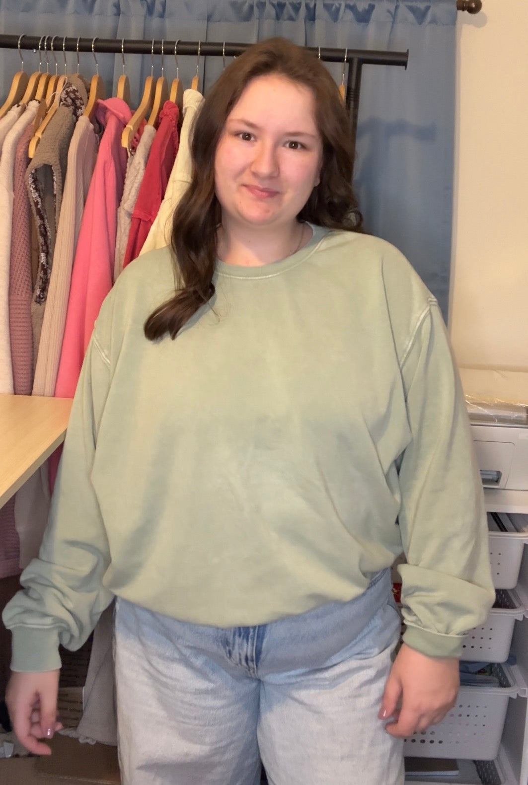 Person wearing a light green sweatshirt and jeans standing in a room with clothes on hangers.