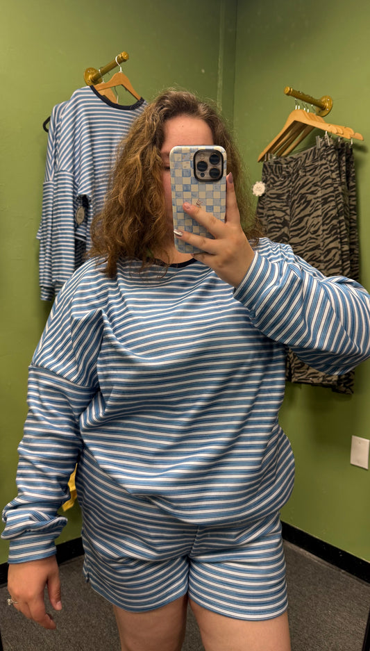 A person taking a mirror selfie in a fitting room wearing a matching blue and white striped long-sleeve top and shorts set. A green wall, wooden hangers, and other clothing are visible in the background.