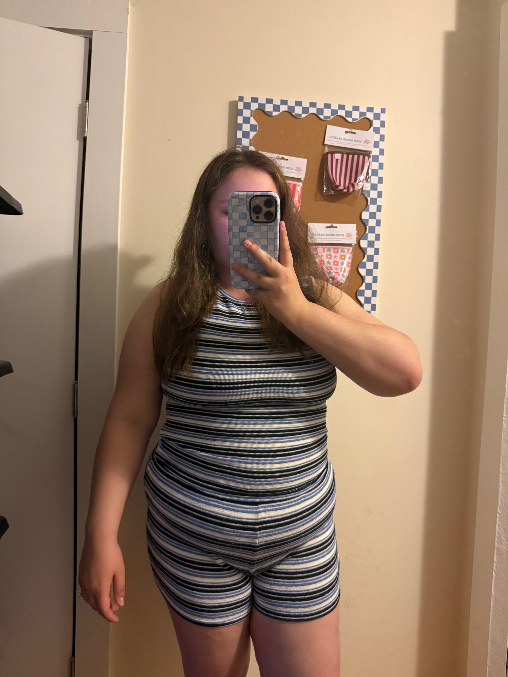 Person taking a mirror selfie wearing a striped outfit in a room with a bulletin board on the wall.