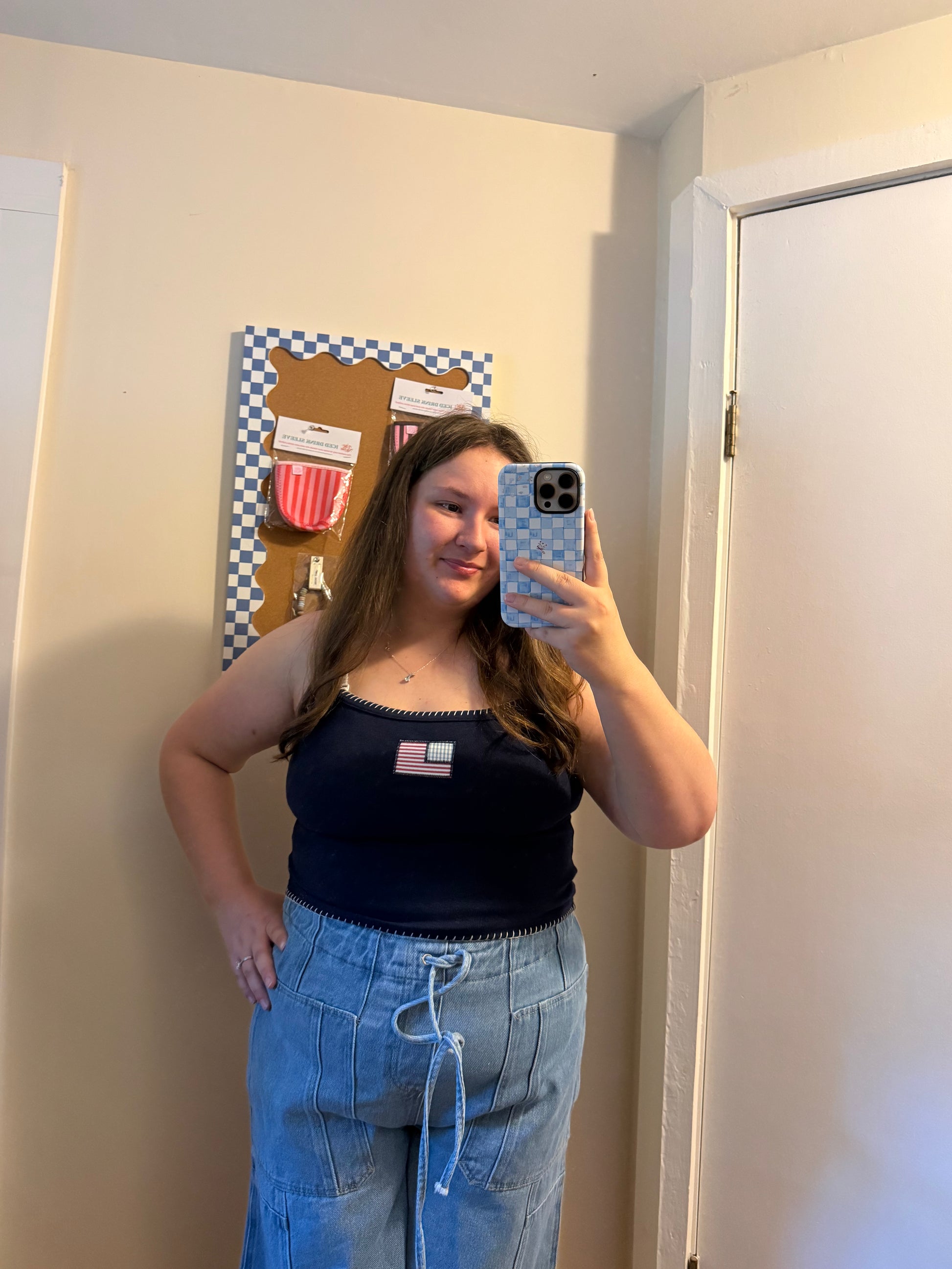 Person taking a mirror selfie wearing a black tank top and blue jeans in a room.