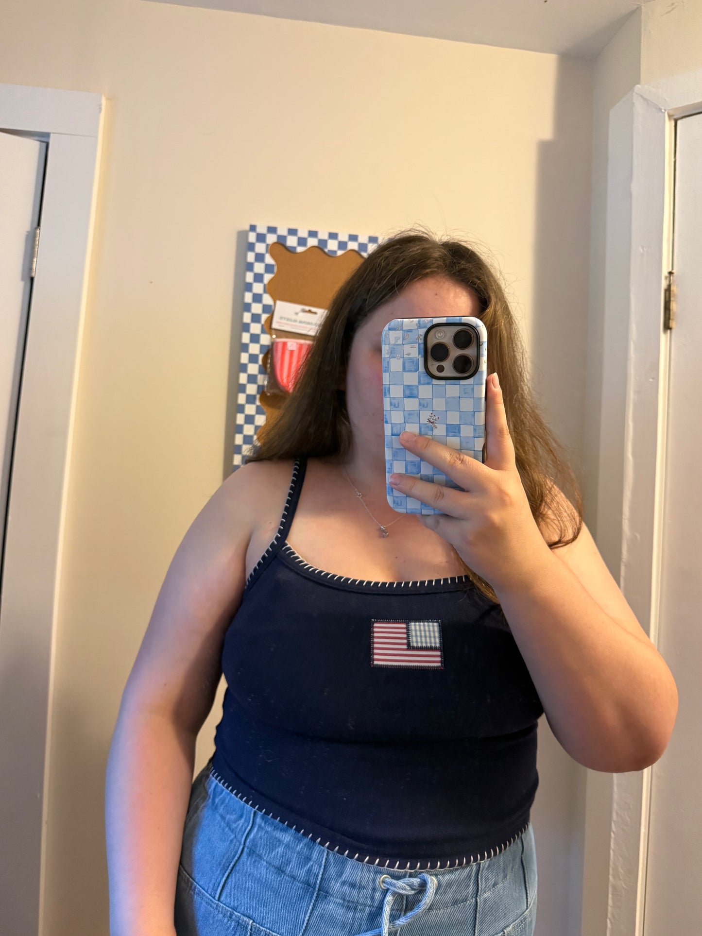 Person taking a mirror selfie wearing a navy tank top with an American flag patch and blue jeans.