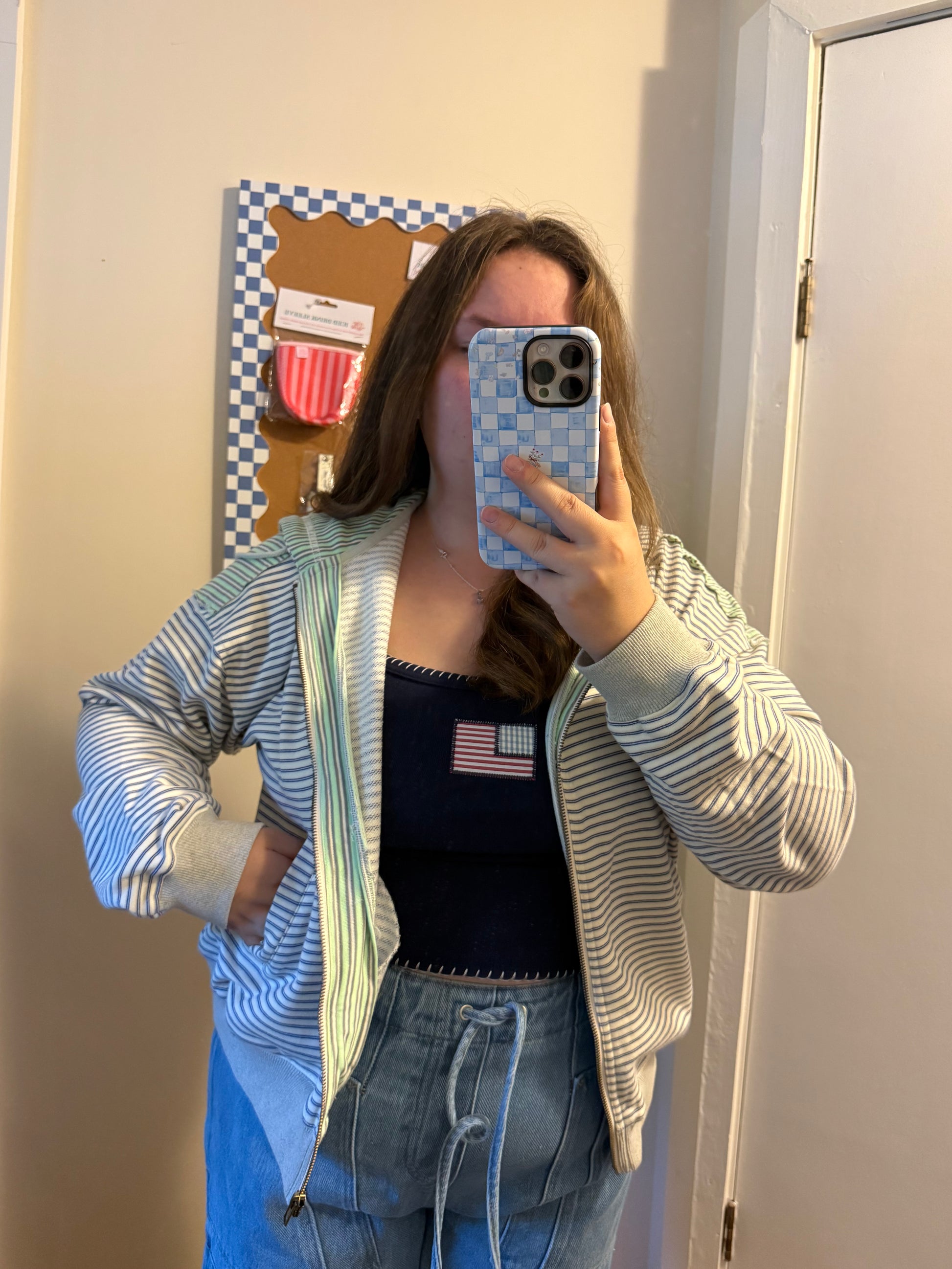 Person taking a mirror selfie wearing a striped hoodie, black top with an American flag, and blue jeans.