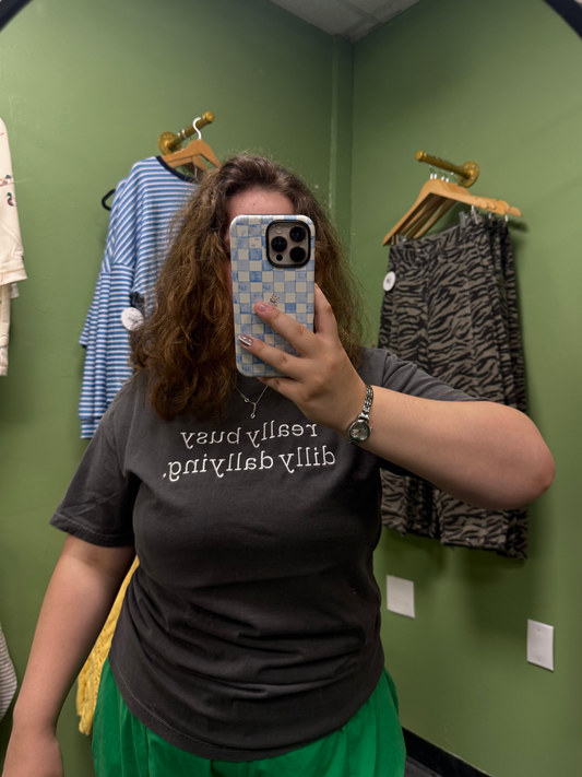 A person taking a mirror selfie in a fitting room wearing a dark gray t-shirt with white text that reads 'dilly dallying really busy' and a bright green skirt. Clothing is hanging on wooden hangers against a green wall in the background.
