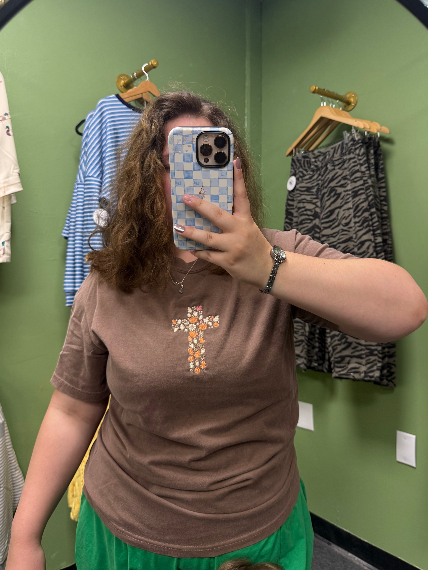 A person taking a mirror selfie in a fitting room wearing a brown short-sleeve t-shirt with a colorful floral cross graphic on the chest and bright green bottoms. Clothing racks with blue and black patterned garments are visible against a green wall.