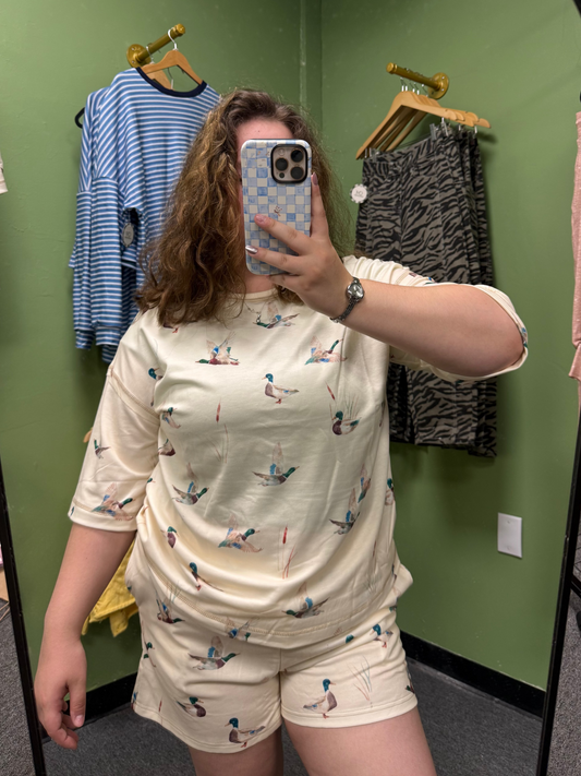 A person taking a mirror selfie in a fitting room wearing a cream-colored two-piece pajama set with a small blue and red bird print. The background shows green walls with other clothes hanging on hangers.