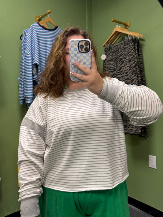 A person taking a mirror selfie in a fitting room wearing a light gray and white striped long-sleeve crop top with gray shoulder patches and a green skirt. Clothing racks with blue and black patterned tops are visible in the green wall background.